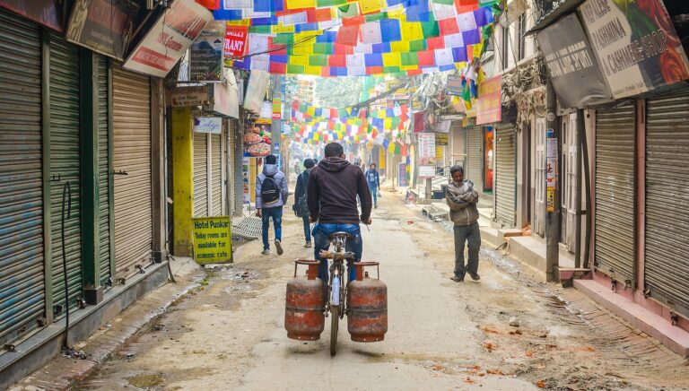 A man riding a bicycle with two large gas cylinders on the carrier down a narrow street lined with closed shops and colorful prayer flags hanging overhead. Another man is walking on the left, and a third man stands on the right, with his arms crossed.