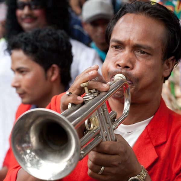 A man in a red jacket playing a trumpet with another man in the foreground and people in costumes visible in the background.