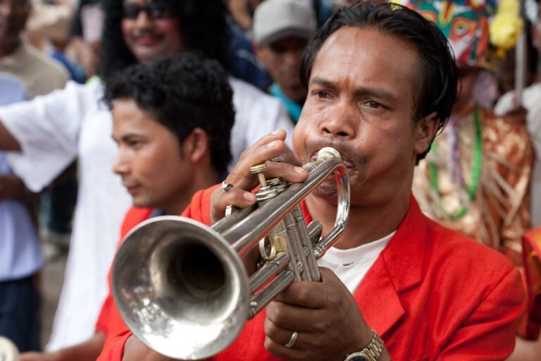 A man in a red jacket playing a trumpet with another man in the foreground and people in costumes visible in the background.