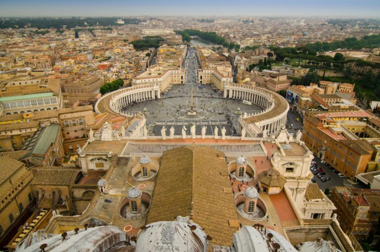 Aerial view of St. Peter's Square in Vatican City, showing the colonnades, central obelisk, and surrounding buildings, with a hazy Rome skyline in the background.