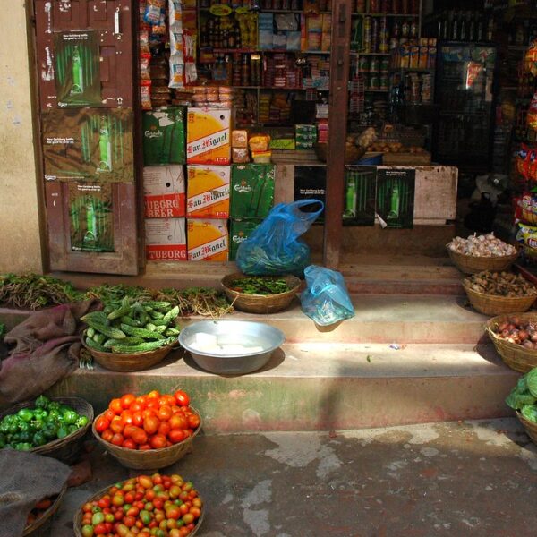 An open-front small grocery store with various fresh vegetables displayed in baskets on the ground in front of packed shelves with food and beverages inside.