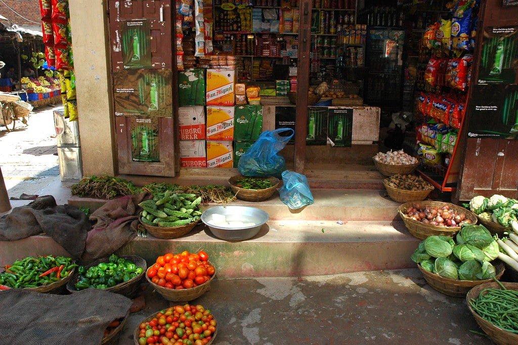 An open-front small grocery store with various fresh vegetables displayed in baskets on the ground in front of packed shelves with food and beverages inside.