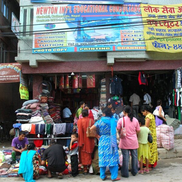 Street scene in Kathmandu, Nepal with pedestrians and shoppers walking past storefronts and street vendors selling clothes and textiles, under signs advertising educational and printing services.