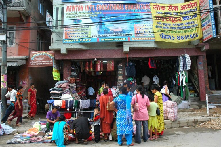 Street scene in Kathmandu, Nepal with pedestrians and shoppers walking past storefronts and street vendors selling clothes and textiles, under signs advertising educational and printing services.