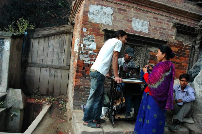 A street scene in Nepal showing three people interacting around a traditional manual sewing machine, with a woman in vibrant traditional clothing speaking to a man who is standing by the machine operated by another man, against the backdrop of a historic brick structure and a wooden door.