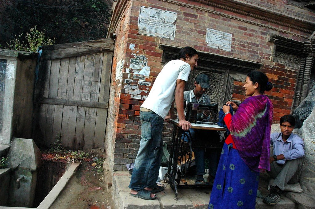 A street scene in Nepal showing three people interacting around a traditional manual sewing machine, with a woman in vibrant traditional clothing speaking to a man who is standing by the machine operated by another man, against the backdrop of a historic brick structure and a wooden door.