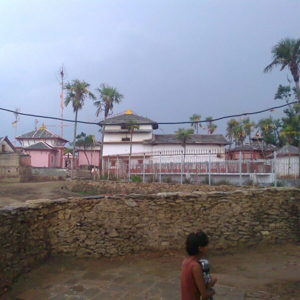 A person standing in front of a walled compound that includes a pink-hued temple with tiered roofs and flag-topped spires, surrounded by various trees under a cloudy sky.