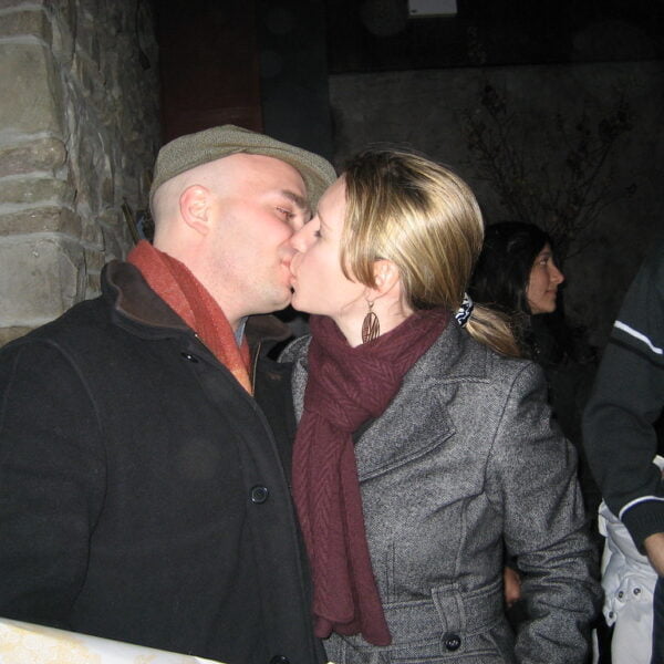 A man wearing a cap and a woman in a coat kissing each other on the lips at an indoor gathering with people in the background.