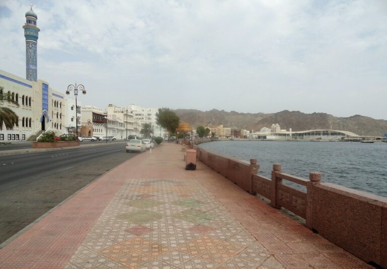 Coastal road with a sidewalk and mosaic tiles, a mosque with a blue patterned minaret, street lamps, and a stone balustrade overlooking a calm sea with hills and buildings in the distance.