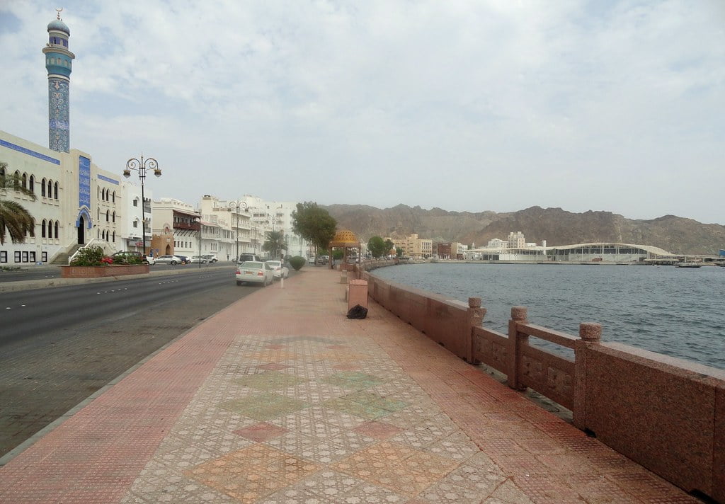 Coastal road with a sidewalk and mosaic tiles, a mosque with a blue patterned minaret, street lamps, and a stone balustrade overlooking a calm sea with hills and buildings in the distance.