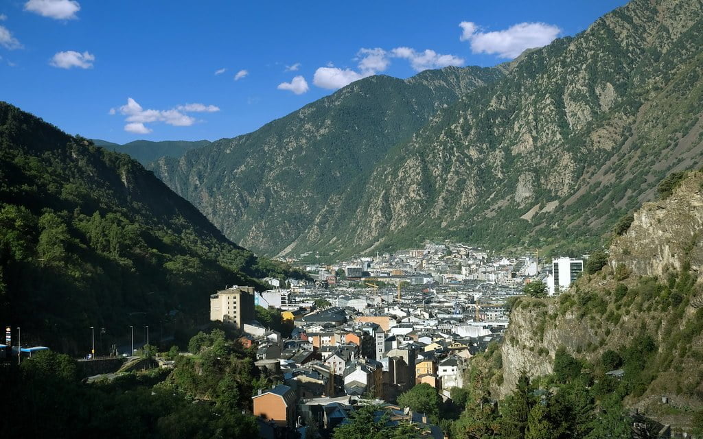 A panoramic view of a town nestled in a mountain valley with dense buildings surrounded by lush, steep hillsides under a clear blue sky.