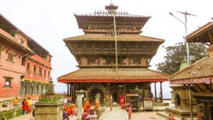 People gathering around a traditional multi-tiered pagoda-style temple with ornate wood carvings in a bustling square, flanked by historic red-brick buildings with tiled roofs.