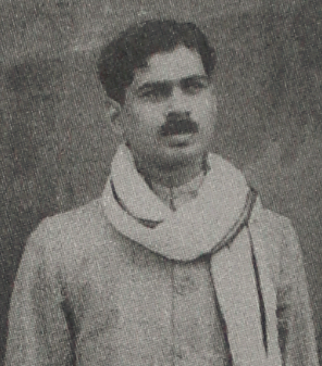 A black and white historical photograph of a man with a mustache, wearing a light-colored coat with a shawl draped around his neck, looking off camera.