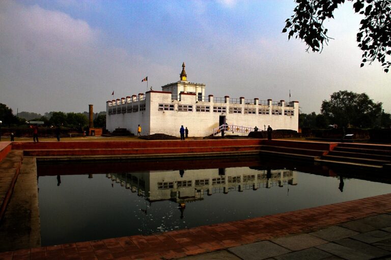 A view of Maya Devi Temple in Lumbini, Nepal, with its white walls and central tower reflected in the still waters of a rectangular pond. People are walking around the temple grounds bordered by trees.