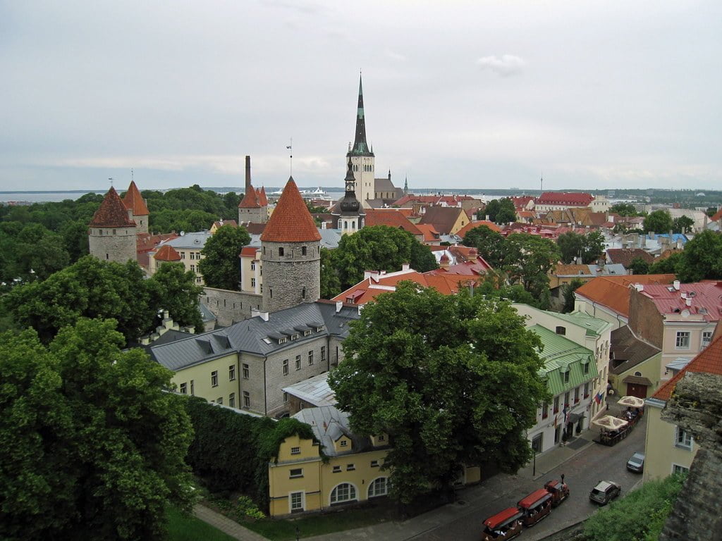 Aerial view of Tallinn's Old Town with medieval towers, historic buildings, and the spire of St. Olaf's Church, under overcast skies with a glimpse of the Baltic Sea in the distance.