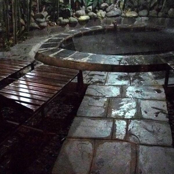 A dimly lit outdoor setting with wooden benches arranged around a circular water feature, stone paving, and a backdrop of dense vegetation.