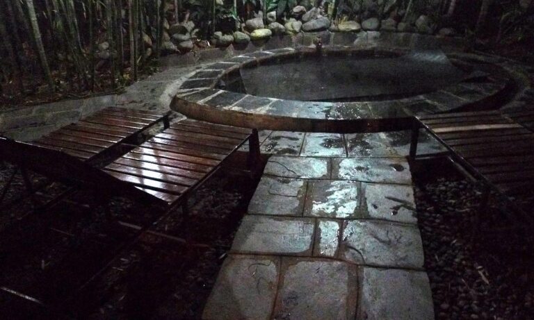 A dimly lit outdoor setting with wooden benches arranged around a circular water feature, stone paving, and a backdrop of dense vegetation.