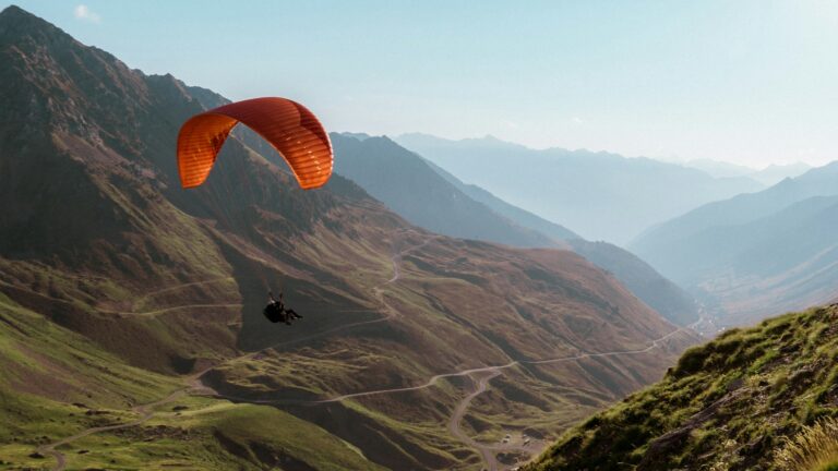 A paraglider flying over a mountainous landscape with winding roads below and a backdrop of mountain ranges.