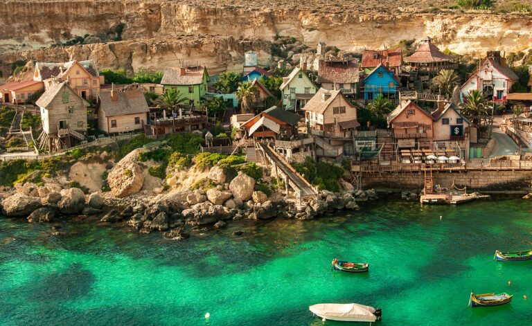 Alt text: A picturesque coastal village with colorful wooden houses built against a cliff above a clear turquoise sea, with small boats moored in the calm water.