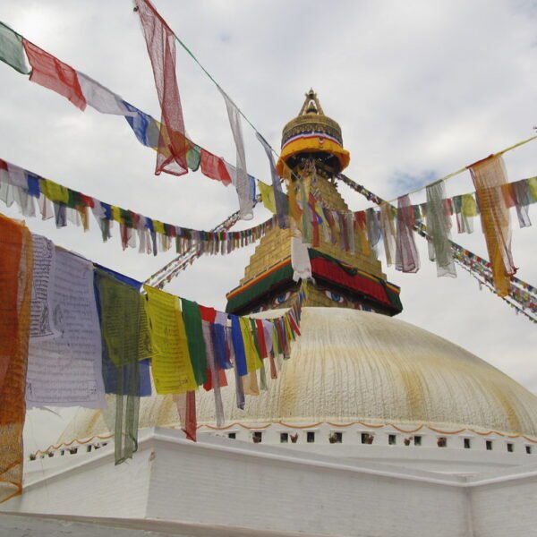 Colorful prayer flags strung across the frame with a large, white Buddhist stupa in the background, featuring a golden spire and eyes painted on it under a cloudy sky.