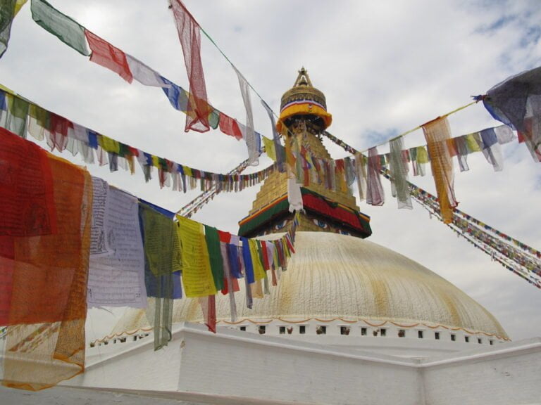 Colorful prayer flags strung across the frame with a large, white Buddhist stupa in the background, featuring a golden spire and eyes painted on it under a cloudy sky.
