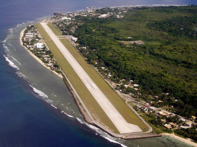 Aerial view of a narrow runway strip on an isthmus with ocean waters on one side and trees and buildings on the other.
