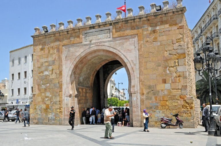 Ancient stone gate with a pedestrian archway in a busy street scene, with people walking and conversing, a parked scooter, street lamps, and a flag on top, under a clear blue sky.