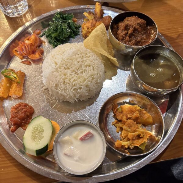 A metal tray with an assortment of Indian dishes including white rice, dal (lentil soup), a spinach dish, pickles, fried appetizers, a raita (yogurt sauce), and what appears to be a dessert, served on a wooden table.
