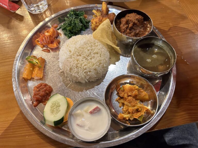 A metal tray with an assortment of Indian dishes including white rice, dal (lentil soup), a spinach dish, pickles, fried appetizers, a raita (yogurt sauce), and what appears to be a dessert, served on a wooden table.