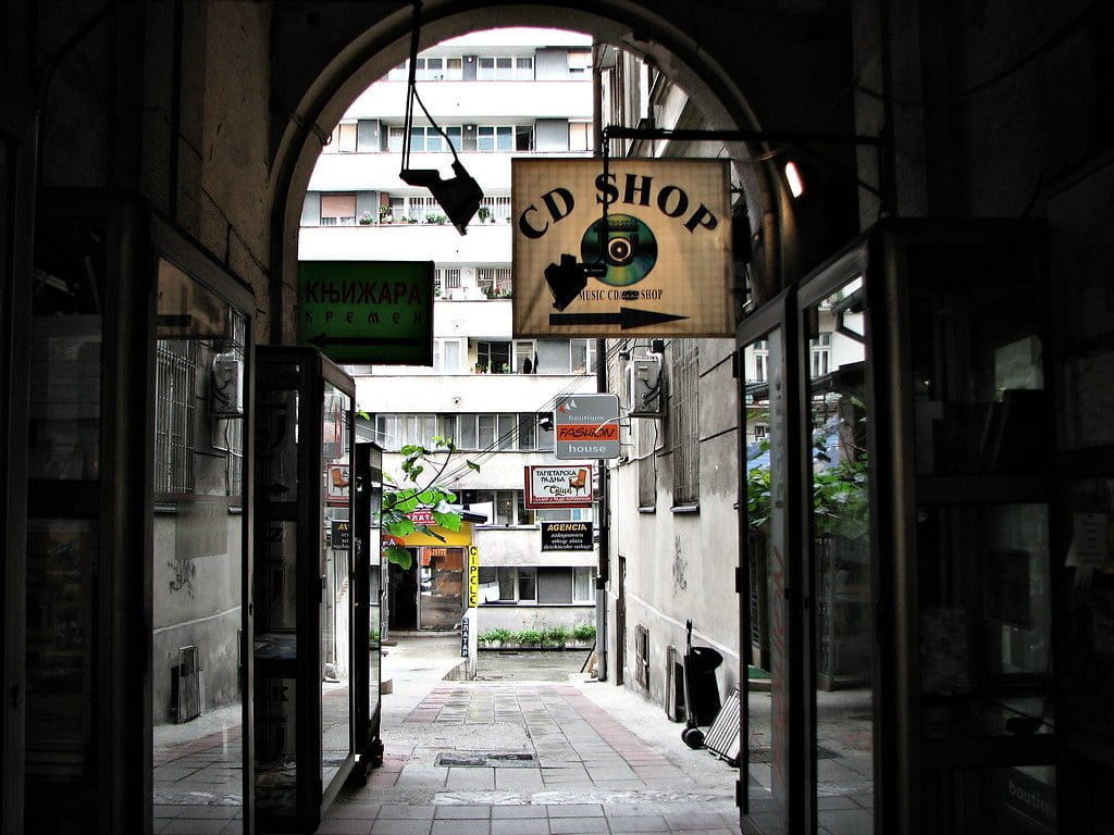 Alt text: An alleyway view with various storefront signs, including a "CD SHOP" sign hanging overhead. The passage leads to an open area with buildings in the background.
