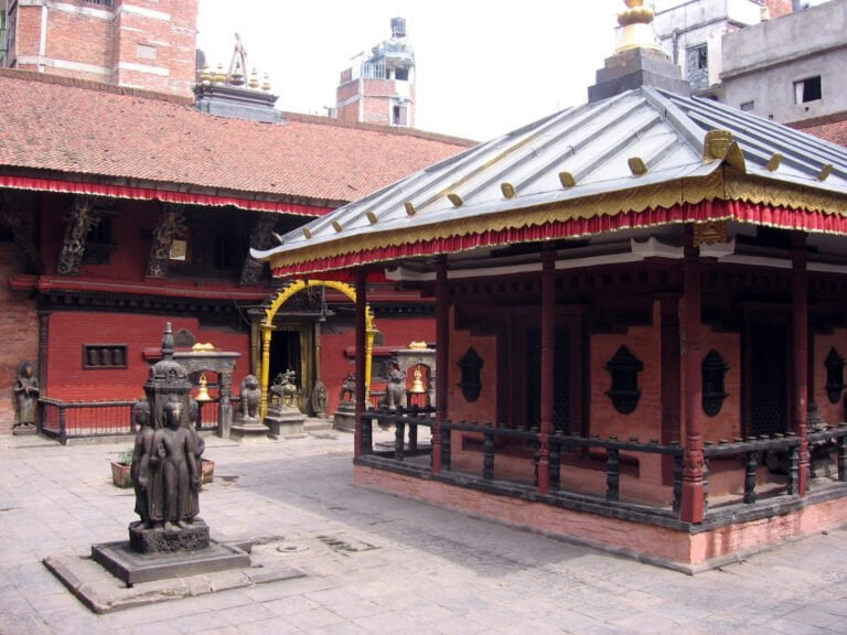 Traditional Nepalese temple complex with red walls, golden details, and multiple statues, including a prominent one in the foreground.