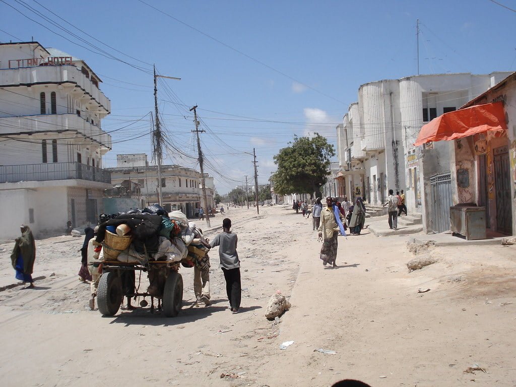 A street scene in a developing country with people walking and a heavily loaded cart being pulled by a donkey, surrounded by buildings and utility poles under a clear sky.