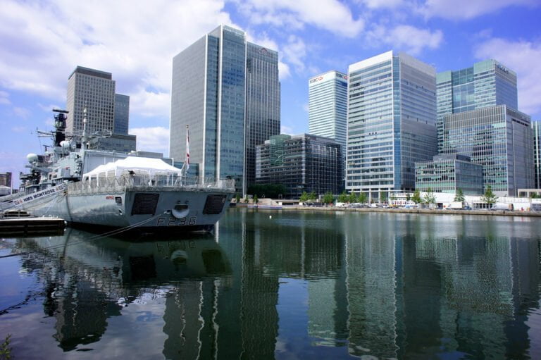 A navy frigate docked in a modern urban waterfront area with a reflection in the water and a backdrop of skyscrapers under a partly cloudy sky.