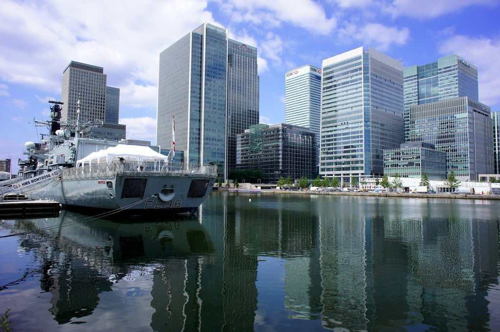 A navy frigate docked in a modern urban waterfront area with a reflection in the water and a backdrop of skyscrapers under a partly cloudy sky.
