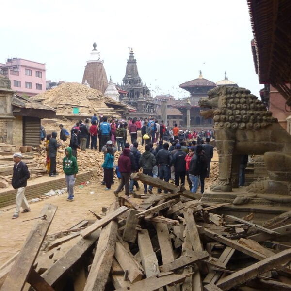 A bustling scene of recovery, where local residents gather amongst wooden debris and damaged structures, possibly after a natural disaster, with an aged stone lion sculpture in the foreground and traditional temples in the background.
