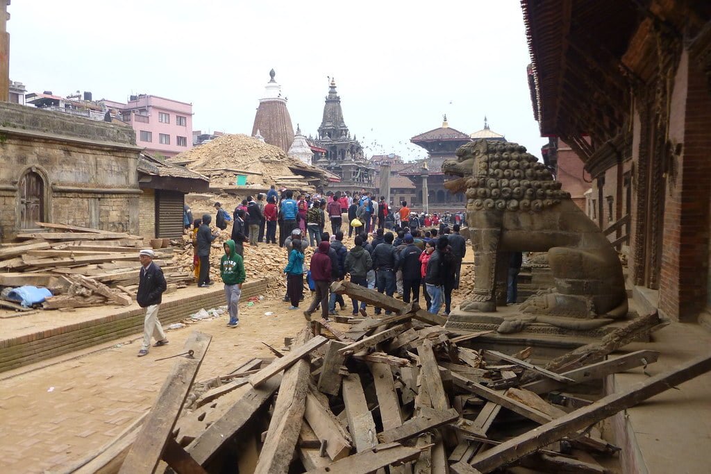 A bustling scene of recovery, where local residents gather amongst wooden debris and damaged structures, possibly after a natural disaster, with an aged stone lion sculpture in the foreground and traditional temples in the background.