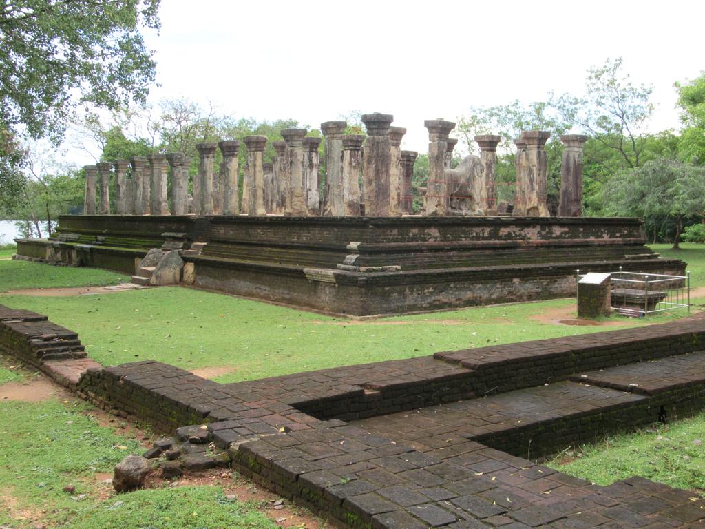 Ancient ruins with rows of stone pillars on a raised platform surrounded by lush greenery, with a dark stone staircase leading up to the structure.