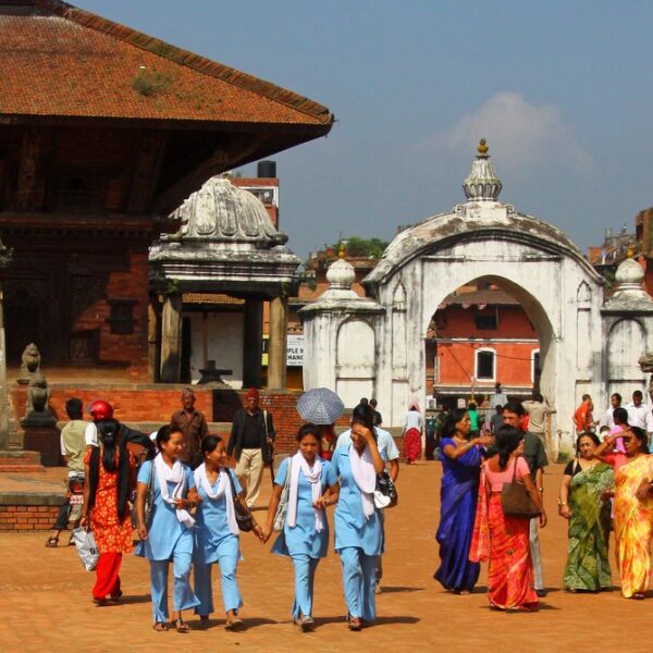 Group of people walking through a historic square with traditional architecture, including a group of students in blue uniforms, near a white arched gateway.