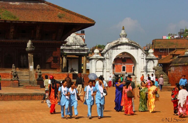 Group of people walking through a historic square with traditional architecture, including a group of students in blue uniforms, near a white arched gateway.