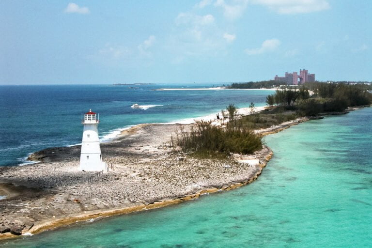 A picturesque white lighthouse on a rocky shoreline with turquoise waters, a sandy beach in the distance, and a large pink hotel complex on the horizon.