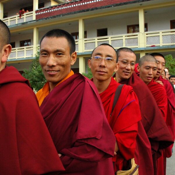 A group of Buddhist monks in traditional red and yellow robes standing in line, with some smiling at the camera, and civilians in the background.