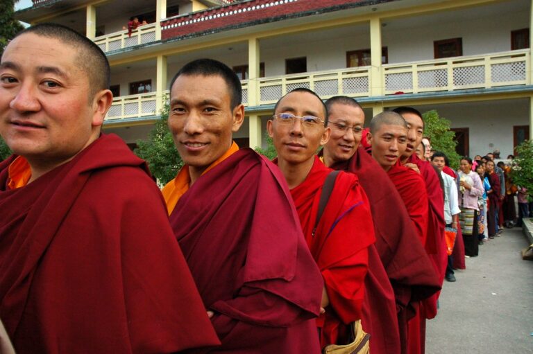 A group of Buddhist monks in traditional red and yellow robes standing in line, with some smiling at the camera, and civilians in the background.