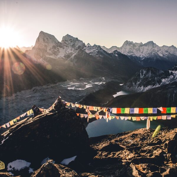 Sunrise over a mountainous landscape with a string of colorful prayer flags in the foreground and snow-capped peaks in the distance. A glacial lake is partially visible in the valley below.