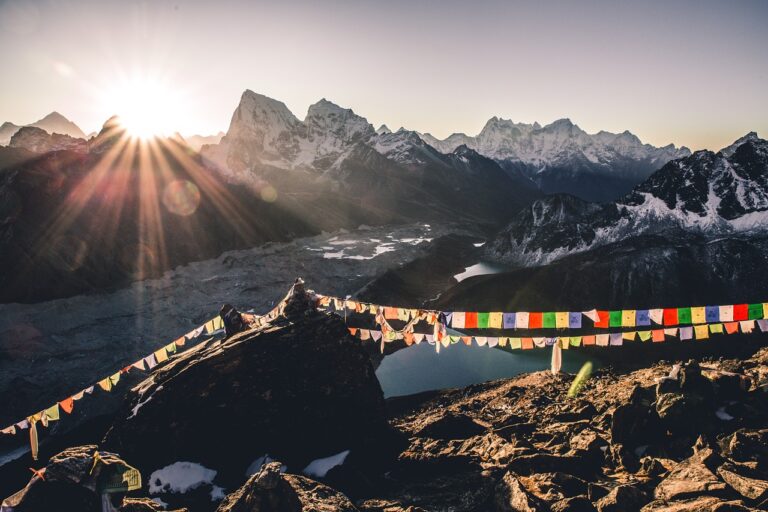 Sunrise over a mountainous landscape with a string of colorful prayer flags in the foreground and snow-capped peaks in the distance. A glacial lake is partially visible in the valley below.