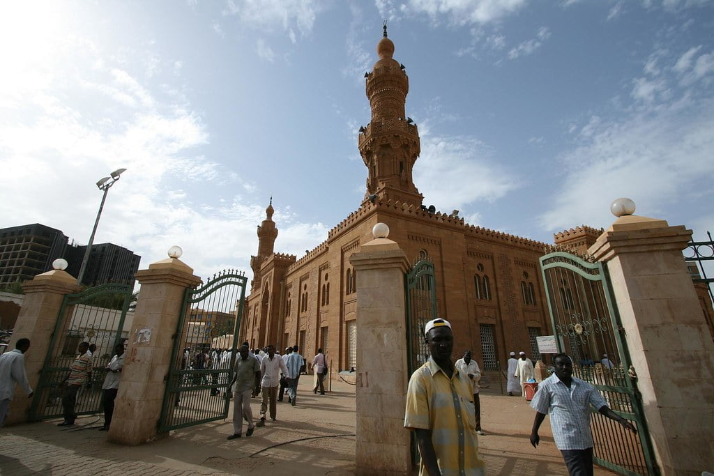 People entering and exiting the gates of a large brick mosque with two minarets under a blue sky.