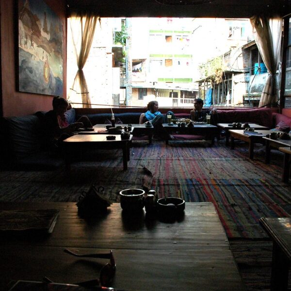 A dimly lit cafe interior with patrons sitting on cushioned benches around low tables, a colorful rug on the floor, and a view of buildings outside through open windows.