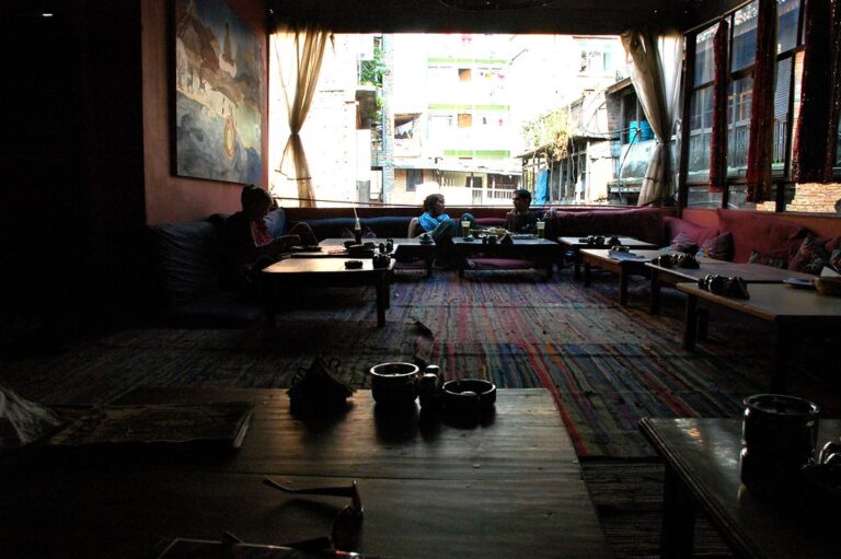A dimly lit cafe interior with patrons sitting on cushioned benches around low tables, a colorful rug on the floor, and a view of buildings outside through open windows.