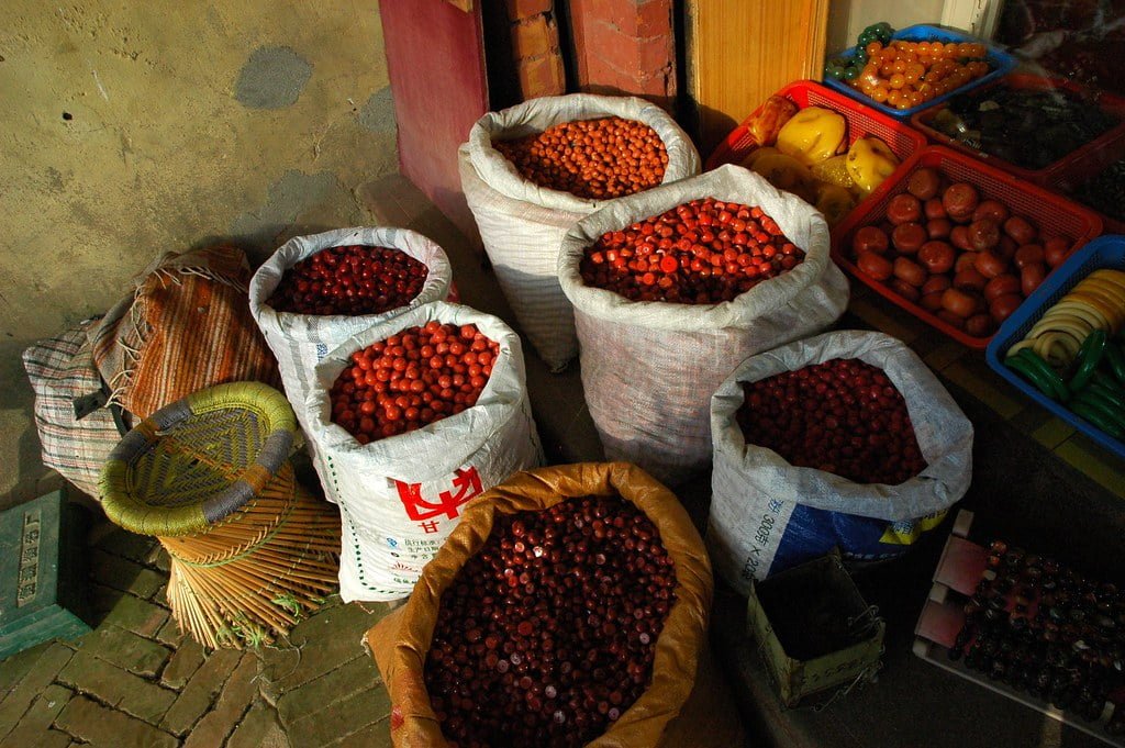 Sacks filled with chestnuts and plastic crates containing various fruits and vegetables displayed in front of a shop with a faded wall in the background.