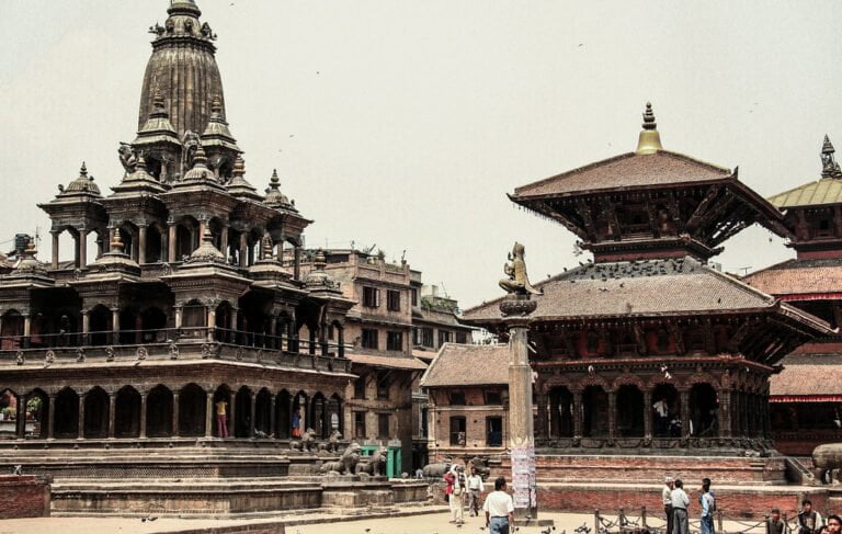 Traditional multi-tiered temples and a statue in the courtyard at Patan Durbar Square in Nepal, with people walking and sitting around the area.
