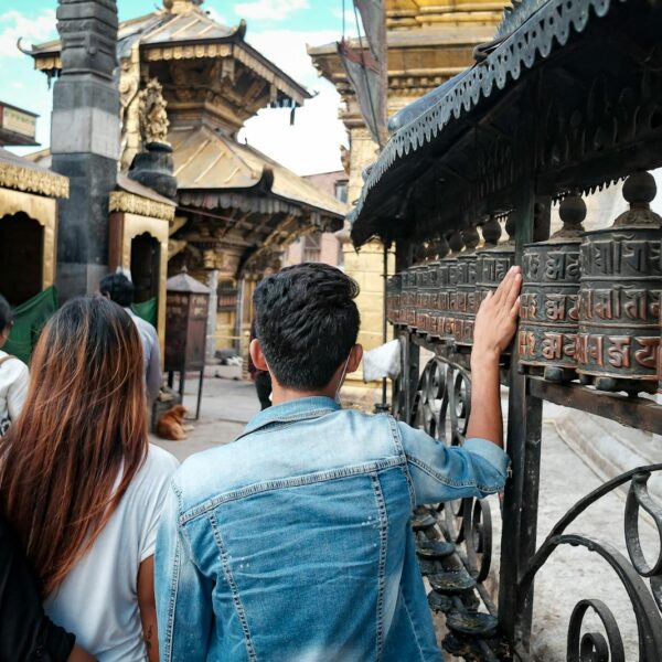 People touching prayer wheels at a temple, with intricate carvings and Nepali script, set against a backdrop of traditional architecture.